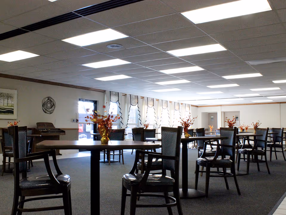A spacious dining room with multiple round tables and chairs arranged neatly. Each table has a small vase with orange and red floral arrangements. The room has large windows with decorative curtains allowing natural light to enter. The ceiling has multiple rectangular fluorescent lights, and there is a piano against one wall.