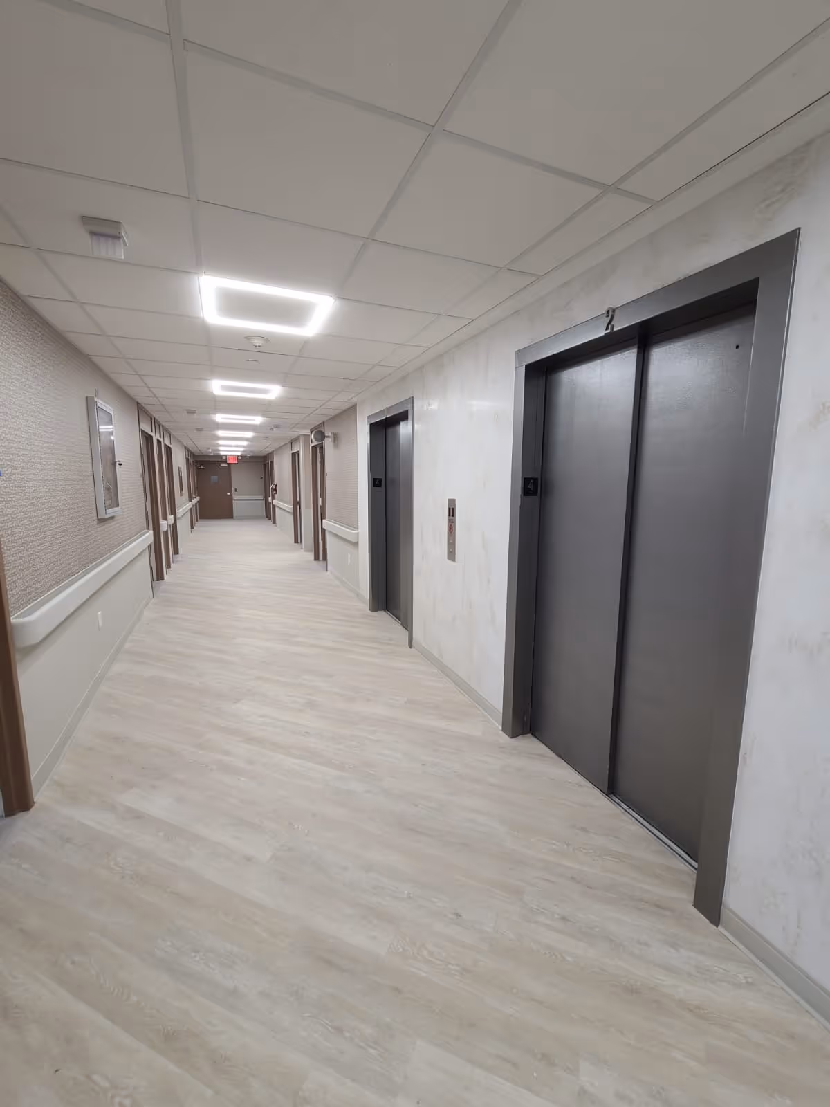Long interior hallway of a care facility with two closed elevators on the right and patient room doors along the corridor.