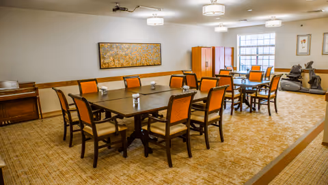 A spacious room with two large dark wood tables surrounded by orange cushioned chairs. The room has beige patterned carpet, light-colored walls, and ceiling lights. There is a large window letting in natural light, exercise equipment in the corner, and a decorative painting on the wall.