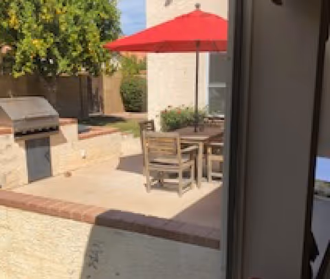 Outdoor patio with a wooden table and chairs under a red umbrella, a built-in grill, and surrounding landscaping.