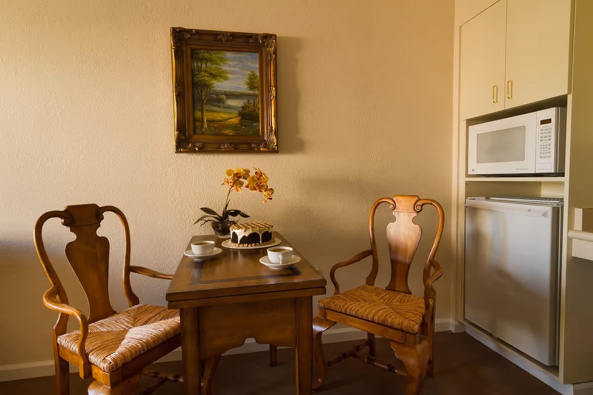 A small dining area with a wooden table and two wooden chairs with woven seats. On the table, there is a cake, two cups with saucers, and a small potted orchid plant. A framed landscape painting hangs on the beige wall above the table. To the right, there is a built-in cabinet with a microwave and a mini refrigerator.