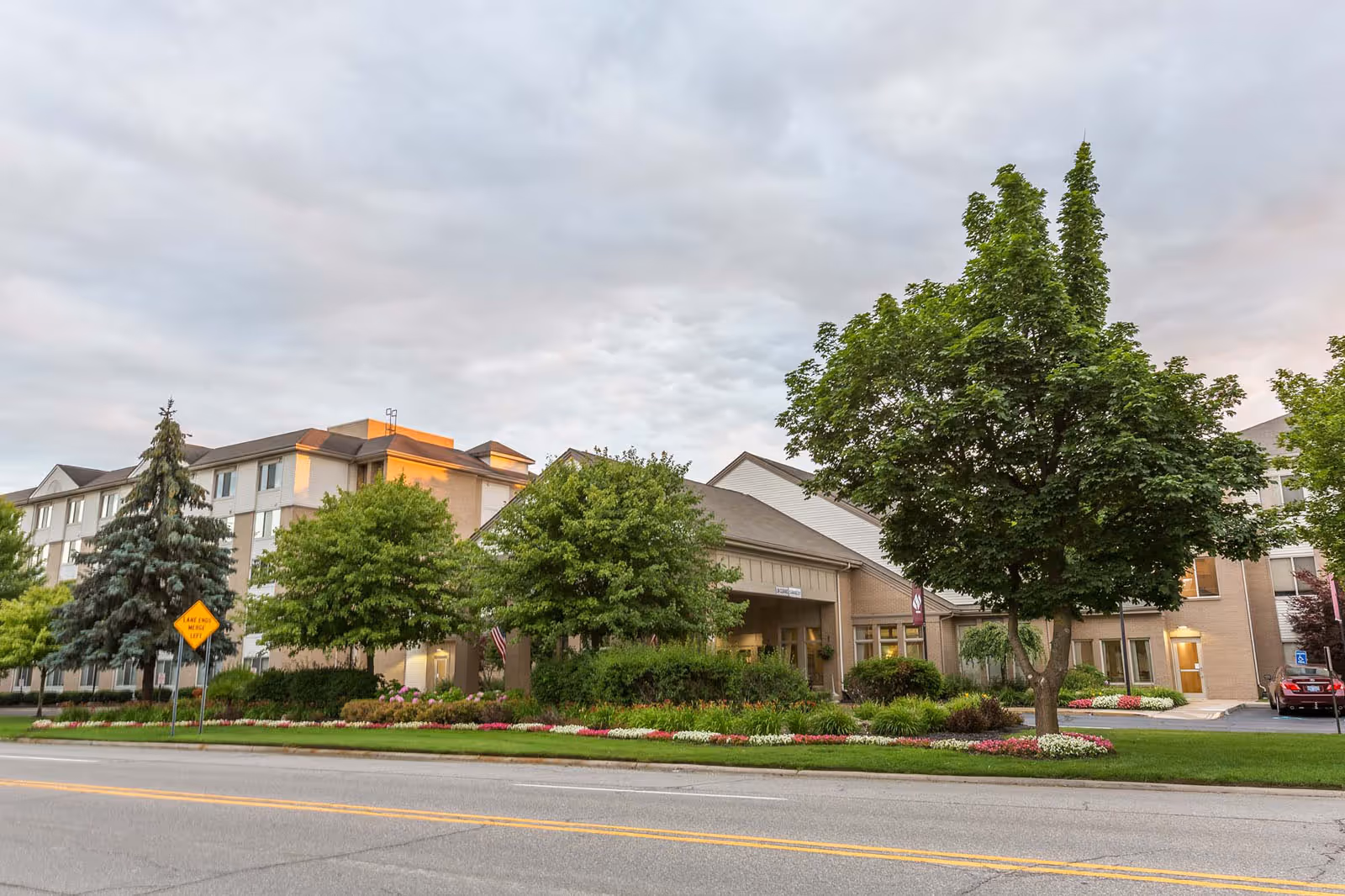 Exterior view of Independence Village of Plymouth, a multi-story senior living facility with well-maintained landscaping including trees, bushes, and flower beds along the front. The building has a beige facade with multiple windows and a covered entrance. A road with a lane ends sign is visible in the foreground under a cloudy sky.