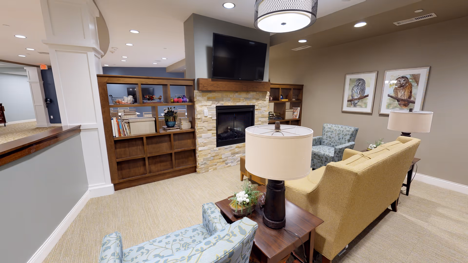 A cozy living room area in an assisted living facility featuring a beige sofa, two patterned armchairs, a wooden side table with a lamp, a stone fireplace with a mounted flat-screen TV above it, and wooden shelving units with books and decorative items. The walls are painted in neutral tones and adorned with framed artwork of owls.