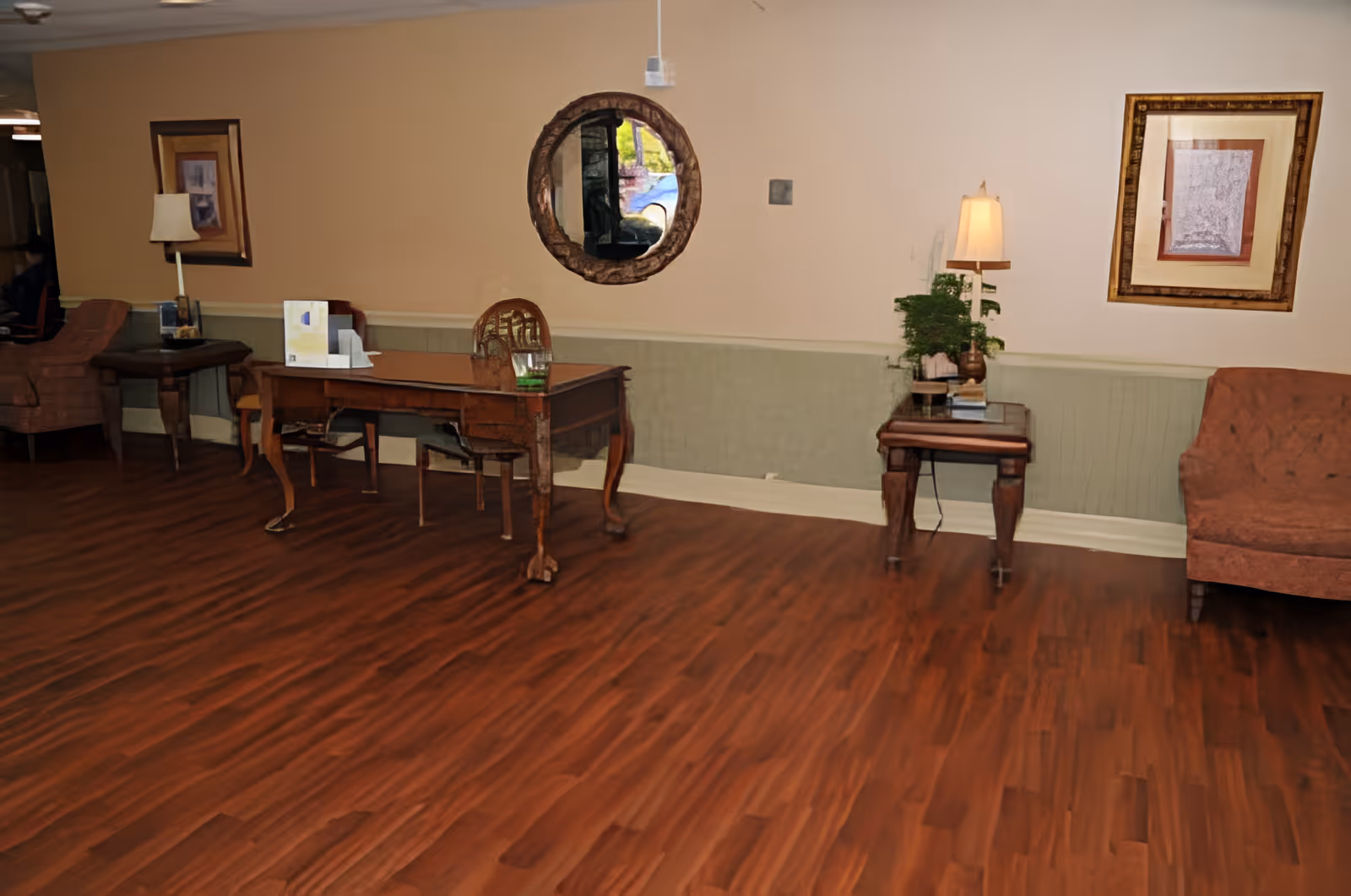 A cozy interior room with wooden flooring, featuring a wooden desk with a chair, two side tables with lamps, a round mirror on the wall, framed artwork, and a cushioned armchair.