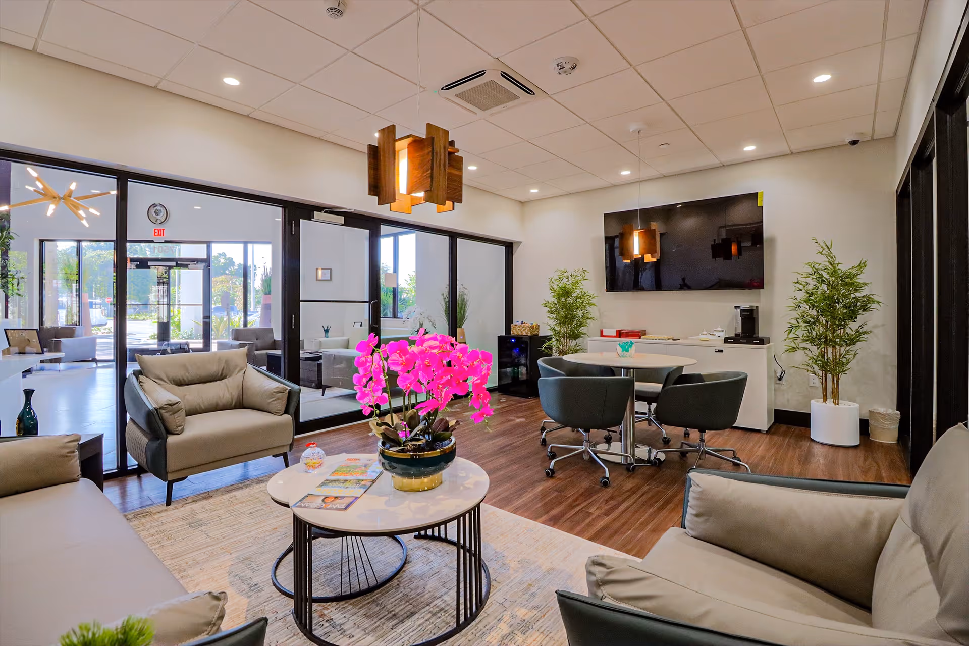 A modern and bright living room area in a senior living facility with beige armchairs and sofas arranged around a white round coffee table with a vibrant pink orchid plant. The room features wooden flooring, a large flat-screen TV mounted on the wall, a round table with four chairs on wheels, and two potted plants. Large glass doors and windows allow natural light to enter, and contemporary wooden pendant lights hang from the ceiling.