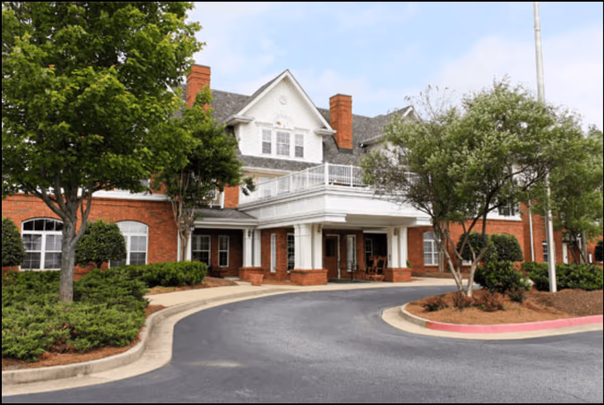 Front exterior view of a senior living facility with red brick walls, white trim, multiple windows, and a covered entrance with columns. There are trees and shrubs around the driveway leading to the entrance.