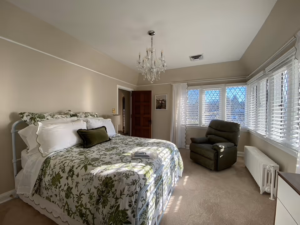 A bright bedroom with a large bed covered in white and green floral bedding, multiple pillows, and a small green cushion. There is a dark green recliner chair near a set of windows with white blinds and curtains. A crystal chandelier hangs from the ceiling, and a wooden door is partially open. The walls are painted beige, and the room has beige carpeting.