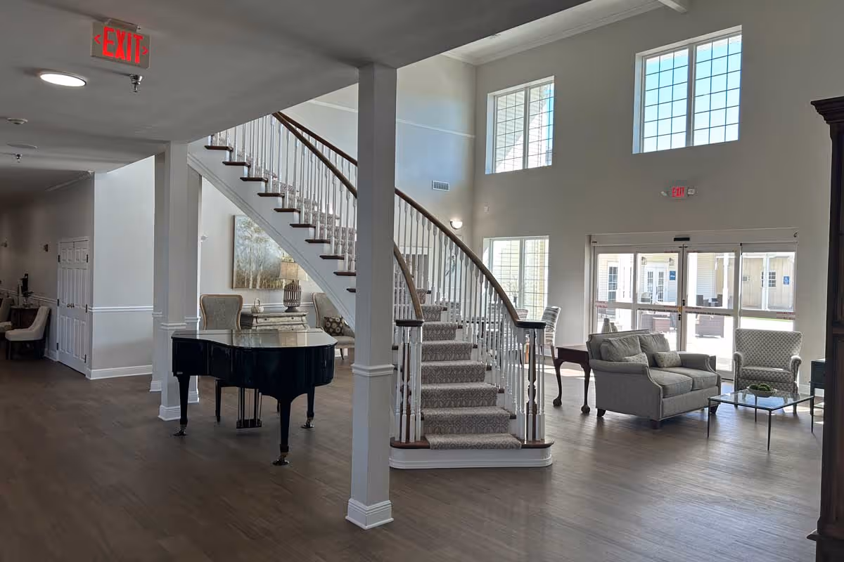 Interior view of a senior living facility lobby with a grand staircase in the center, a black grand piano to the left, and a seating area with a sofa and armchairs near large windows letting in natural light.