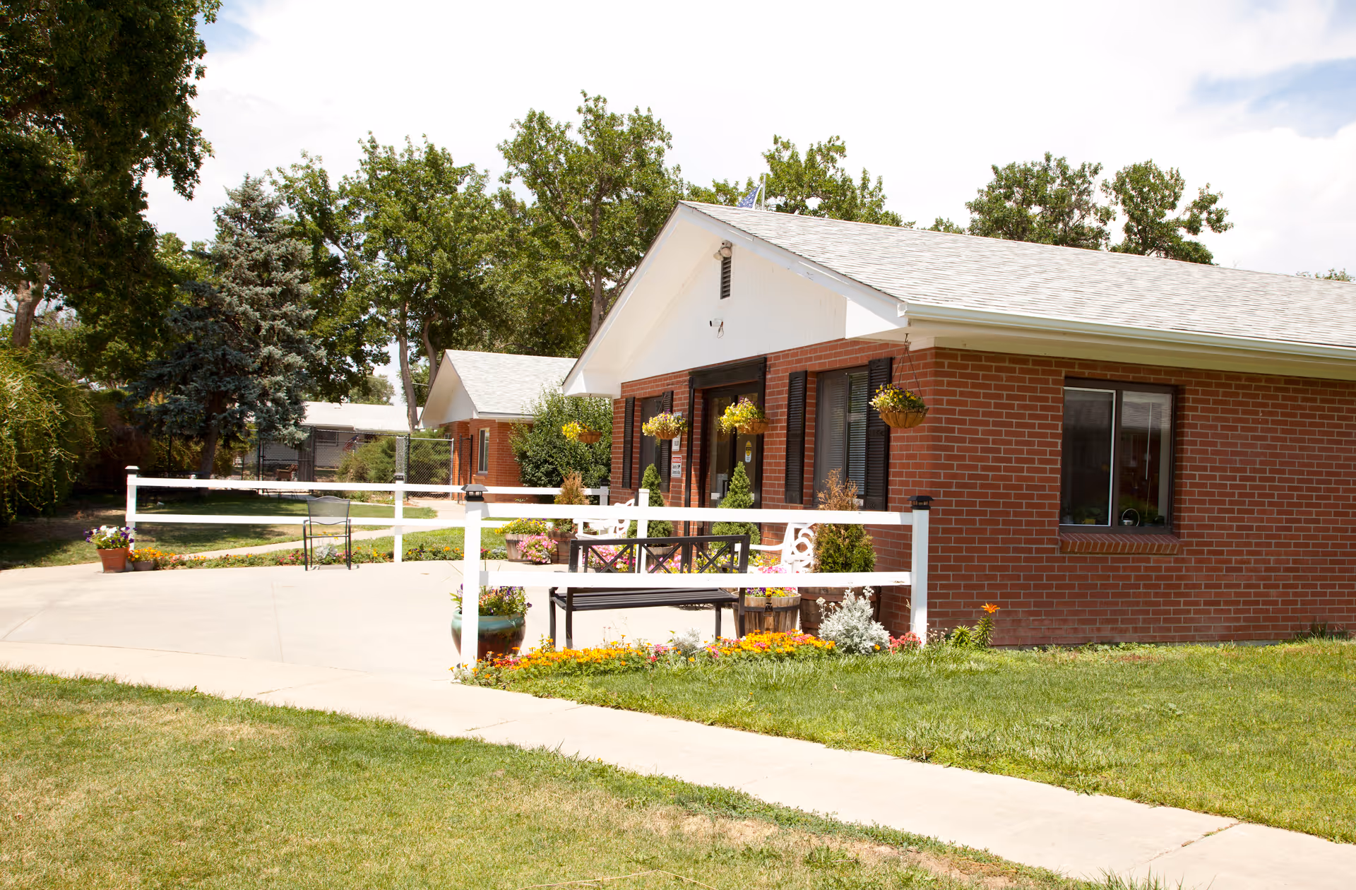 Front exterior of a single-story brick building with a small porch, white fence, hanging flower baskets, benches and landscaped lawn.