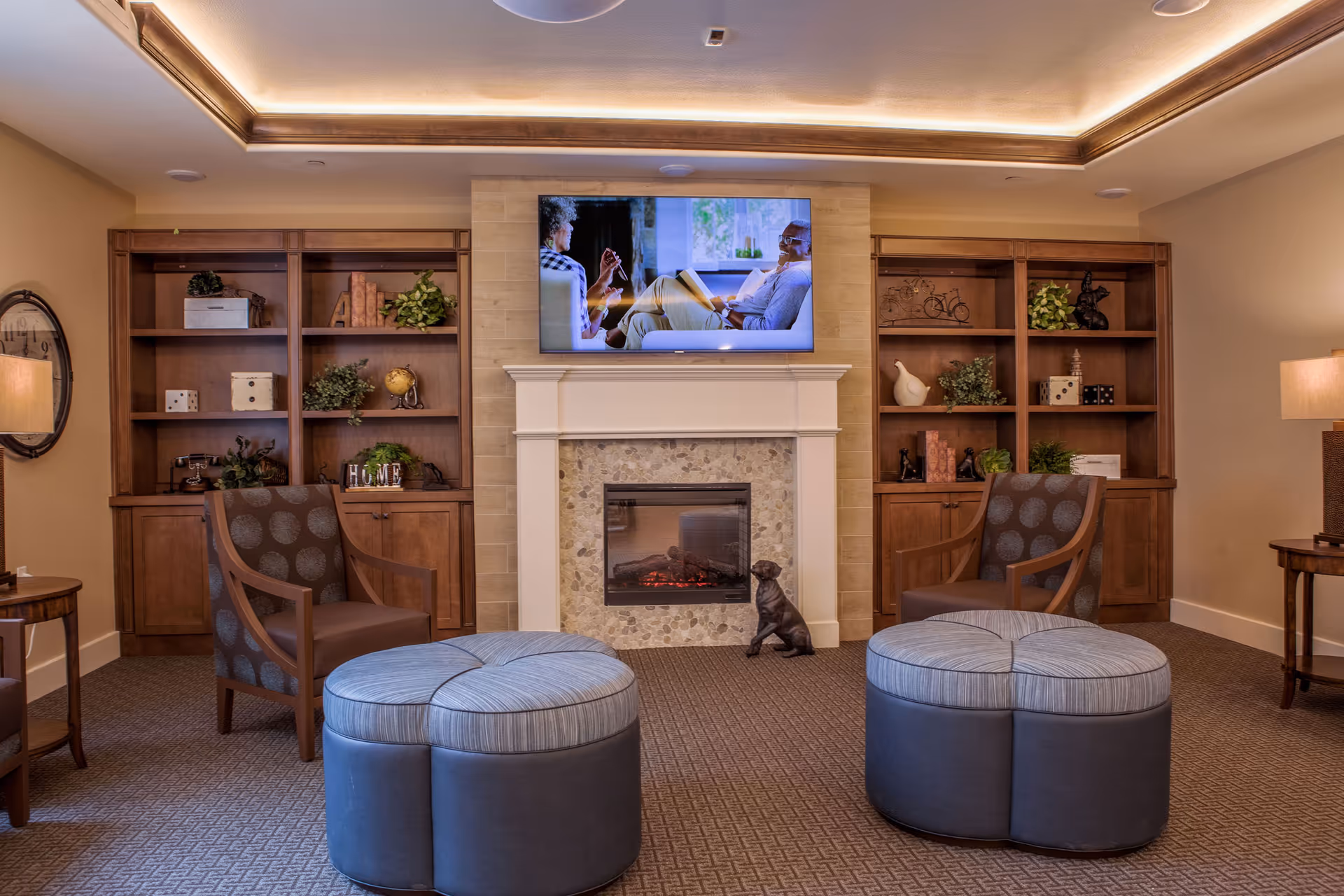 A cozy senior living room with two patterned armchairs, two round ottomans, wooden bookshelves filled with decorative items, a fireplace with a stone surround, and a flat-screen TV mounted above the fireplace displaying two people conversing.