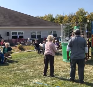 An outdoor gathering at Hearthstone Village with elderly people sitting on chairs and socializing on a grassy lawn. A woman is preparing to throw a ball at a dunk tank where a person is seated inside. The background shows a single-story building with windows and a banner.