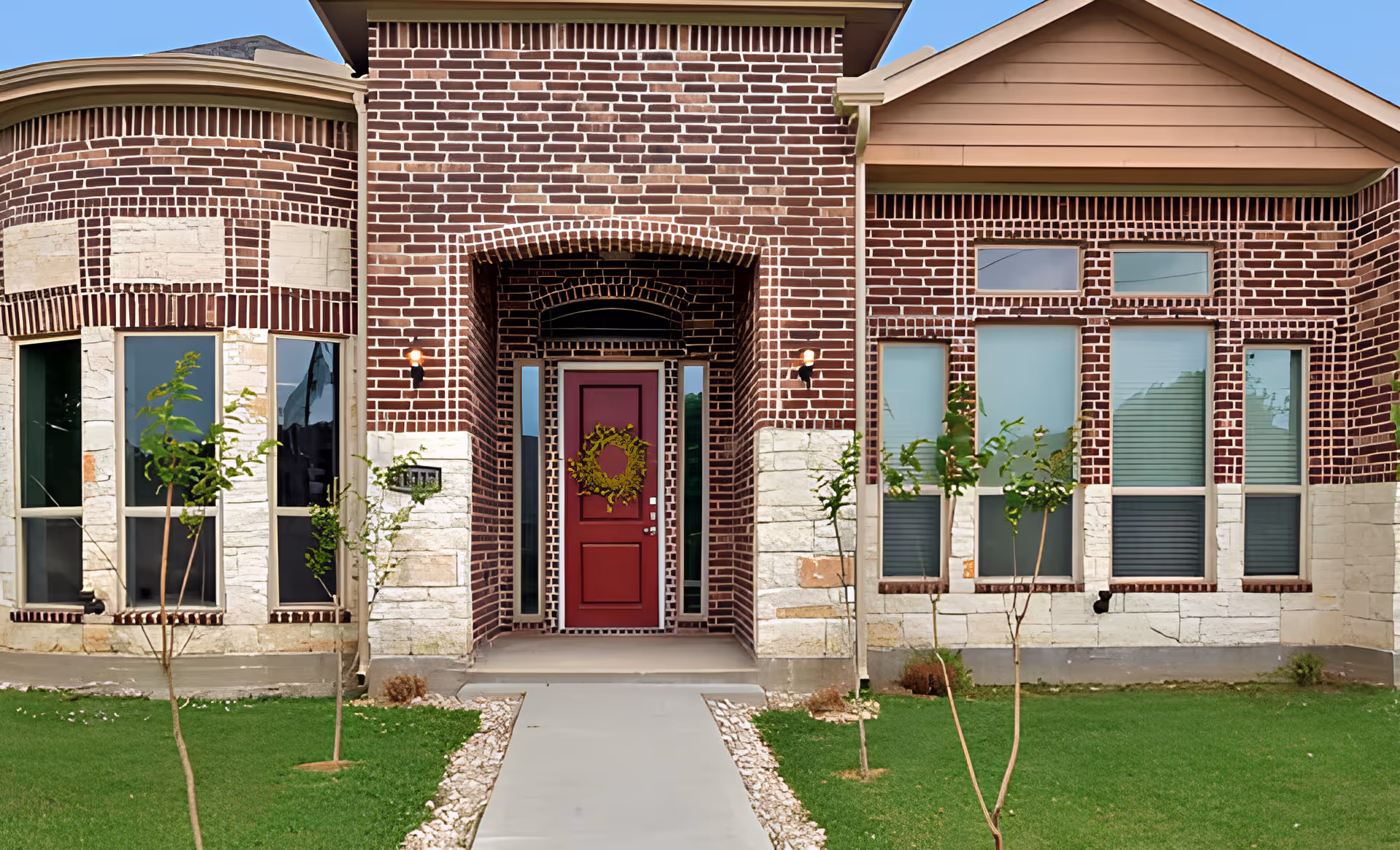 Front exterior view of a brick building with a red door decorated with a wreath, flanked by tall windows and small trees planted in a green lawn.