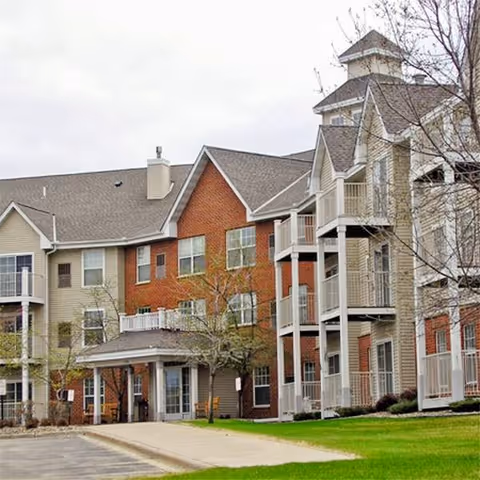 Exterior view of a multi-story senior living facility named Trinity Terrace with a combination of brick and beige siding, multiple balconies, a covered entrance, and a well-maintained lawn and trees in front.