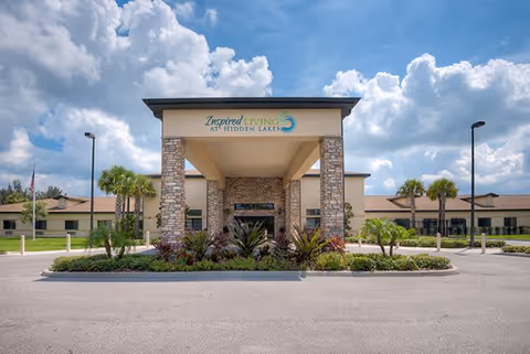 Front exterior view of the Inspired Living Hidden Lakes facility with a covered entrance supported by stone pillars, landscaped greenery, palm trees, and a partly cloudy sky in the background.