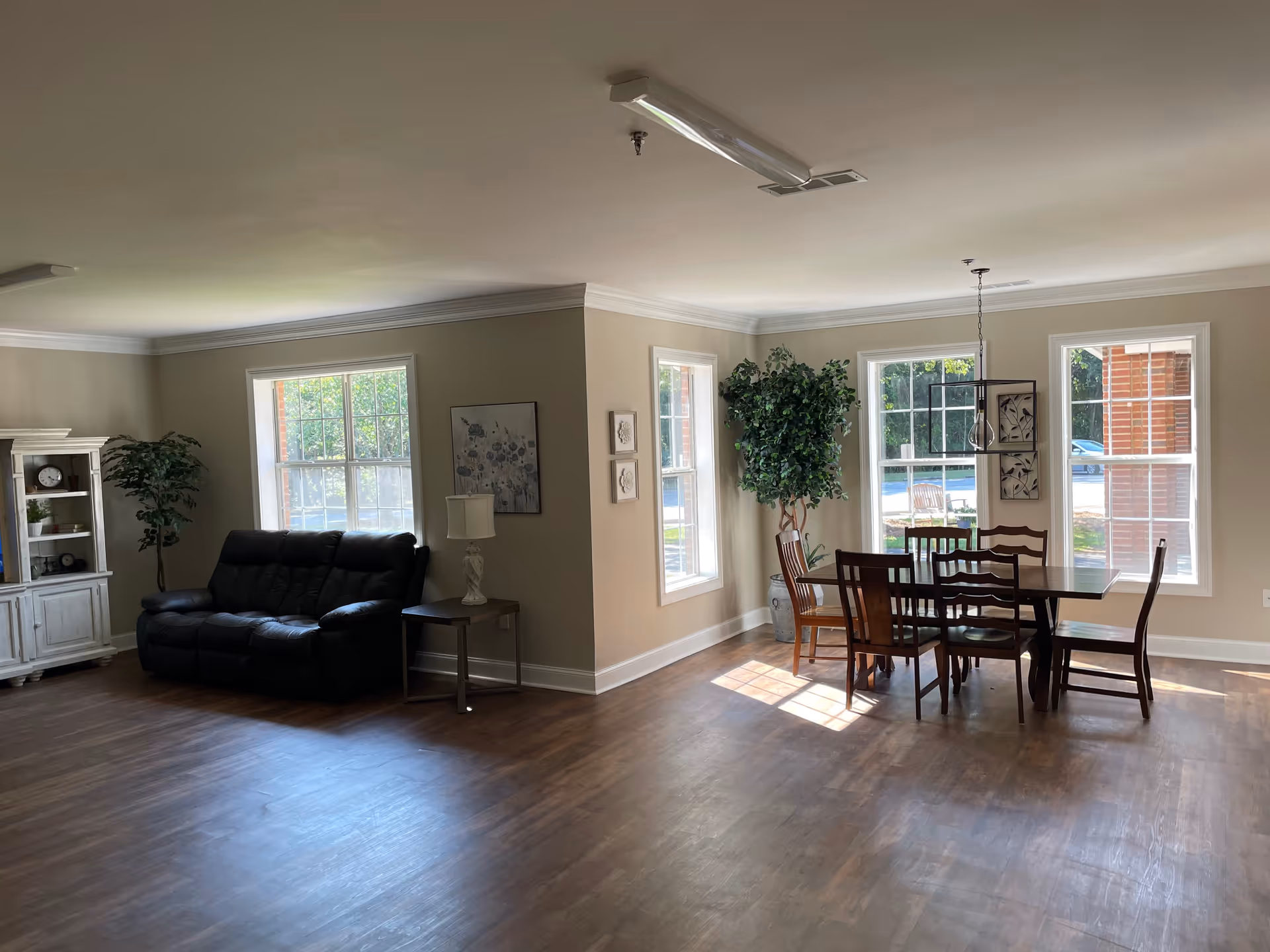 A bright room with wooden flooring featuring a black leather sofa, a white wooden cabinet with decorative items, a side table with a lamp, and a dining area with a wooden table and six chairs near large windows letting in natural light. There are also two large potted plants and framed artwork on the walls.