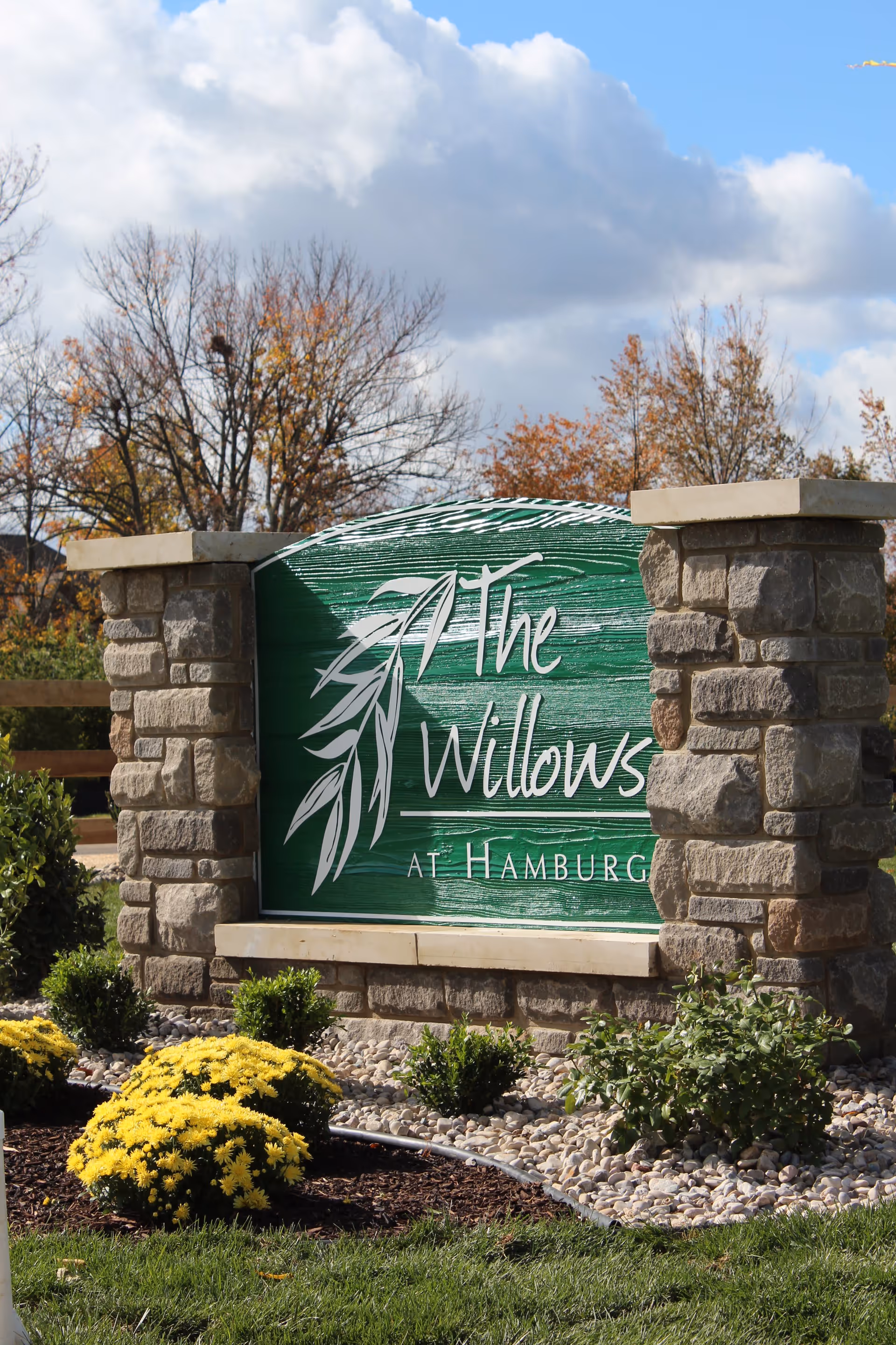 Stone signpost with a green wooden sign that reads 'The Willows At Hamburg' surrounded by landscaping including yellow flowers, bushes, and rocks, with trees and a partly cloudy sky in the background.