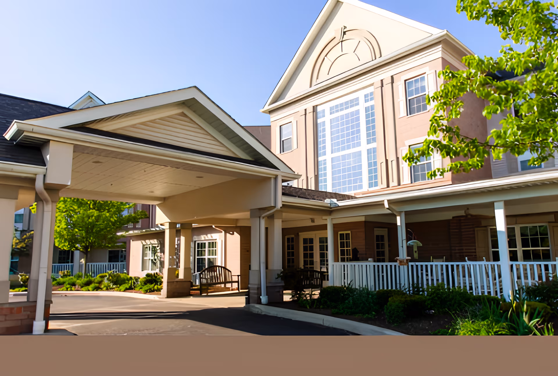 Exterior view of a senior living facility building with a covered entrance driveway, large windows, and a porch area with benches and white railings, surrounded by greenery and trees under a clear blue sky.