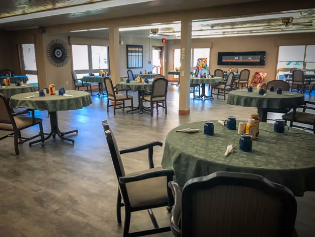 A spacious dining room with multiple round tables covered in green tablecloths. Each table has several chairs around it and is set with blue mugs, napkins, and condiment bottles. The room has large windows allowing natural light to enter, and there are decorative elements on the walls.