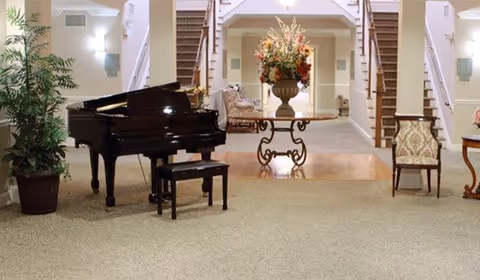 Interior view of a senior living facility lobby with a black grand piano and bench on the left, a large potted plant beside it, and a decorative table with a large floral arrangement in the center. Two staircases with wooden railings lead up on either side of the room. There are upholstered chairs and soft lighting on the walls.