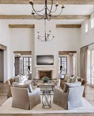 A spacious and elegant living room with high ceilings and exposed wooden beams. The room features two beige sofas and four matching armchairs arranged around a wooden coffee table on a light-colored rug. A fireplace with a mounted flat-screen TV above it is centered on the far wall, flanked by two large windows with curtains. Two black chandeliers hang from the ceiling, adding a touch of sophistication to the bright and airy space.