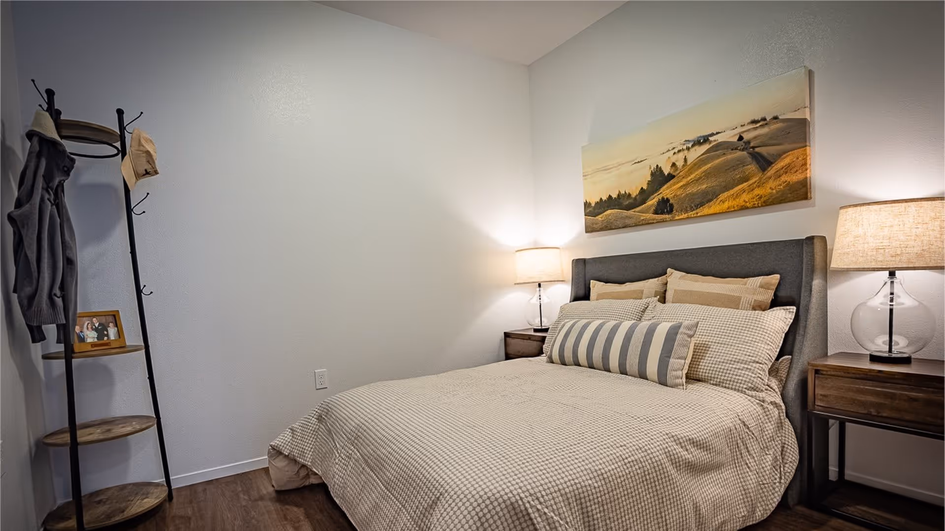 Bedroom with a made bed featuring a striped bolster pillow, two nightstands with lamps, wall art above the headboard, and a corner coat rack.