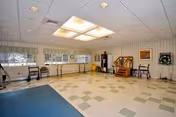 A spacious rehabilitation room with a checkered tile floor, large windows along one wall, and ceiling lights. The room contains exercise equipment including parallel bars, a small set of stairs with a handrail, chairs, and a dartboard on the wall.