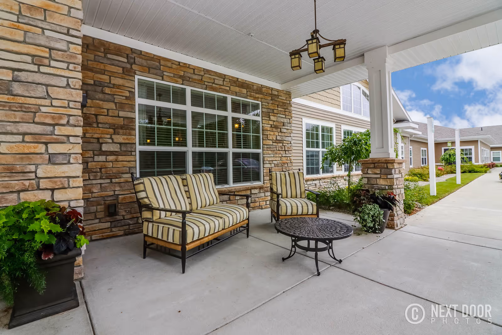 Covered outdoor patio area with stone and siding exterior walls, featuring a cushioned striped loveseat and armchair, a round metal coffee table, potted plants, and a hanging light fixture under a white ceiling. The patio overlooks a driveway and landscaped greenery under a partly cloudy sky.