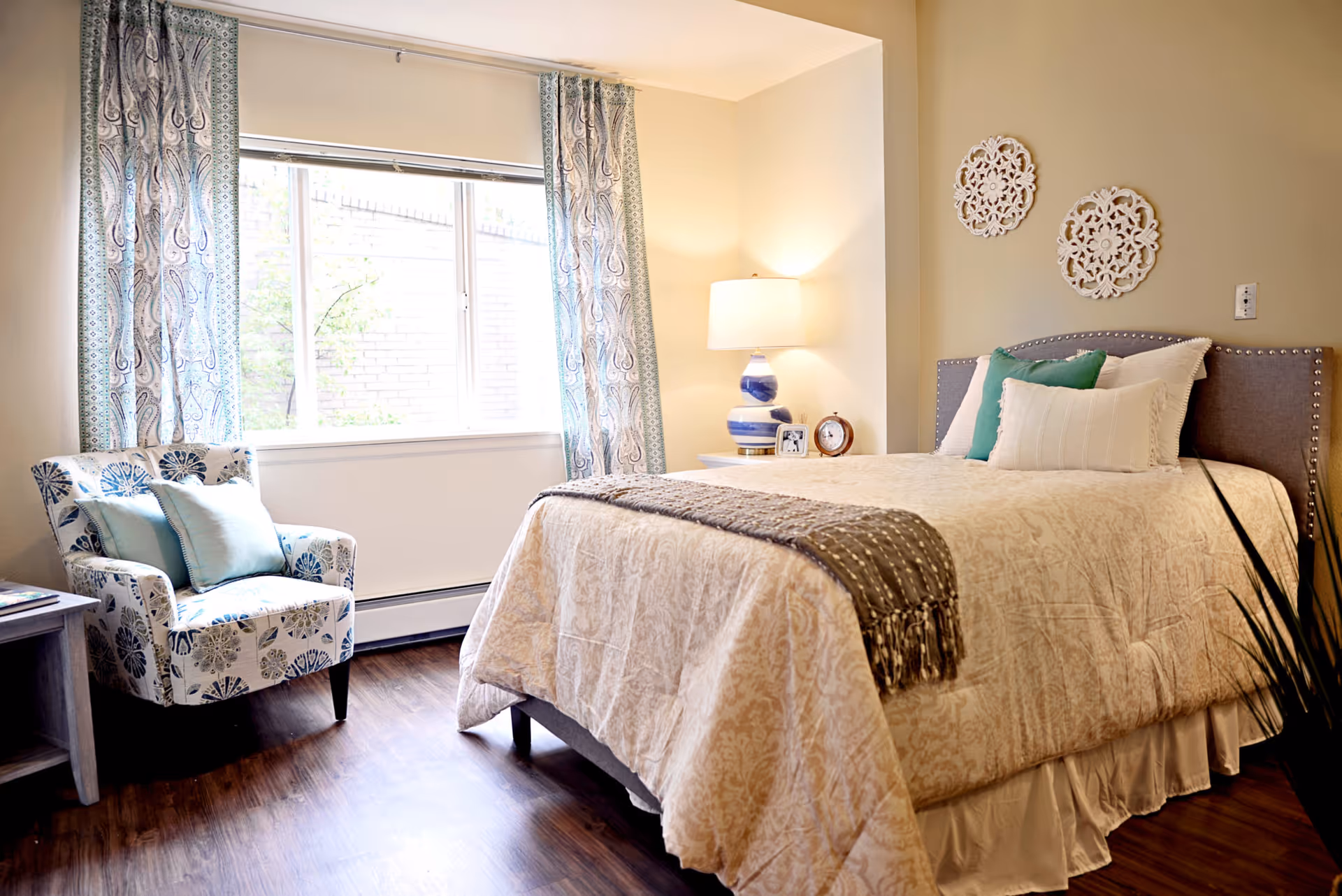 A cozy bedroom with a neatly made bed featuring beige patterned bedding and multiple pillows. There is a bedside table with a blue and white lamp, a clock, and a framed photo. A patterned armchair with blue and beige cushions sits near a window with patterned curtains. The room has wooden flooring and light-colored walls with decorative wall hangings above the bed.