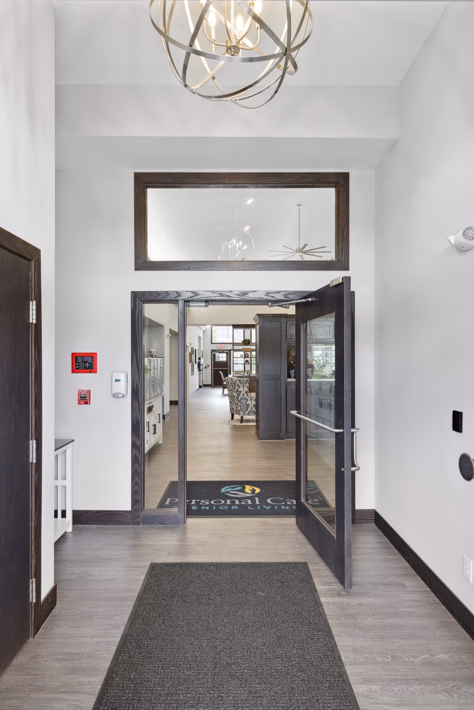 View of an entrance hallway in a senior living facility with an open glass door leading to a common area furnished with chairs and cabinets. The floor has a dark mat with the text 'Personal Care Senior Living'. The walls are white with dark wood trim, and a modern spherical chandelier hangs from the ceiling.