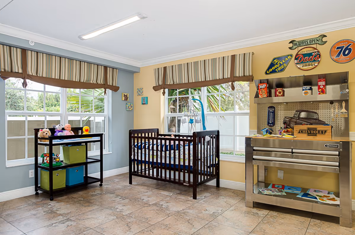 Bright nursery-style room with a crib, toy storage shelves, and a metal tool-bench play area beneath large windows.