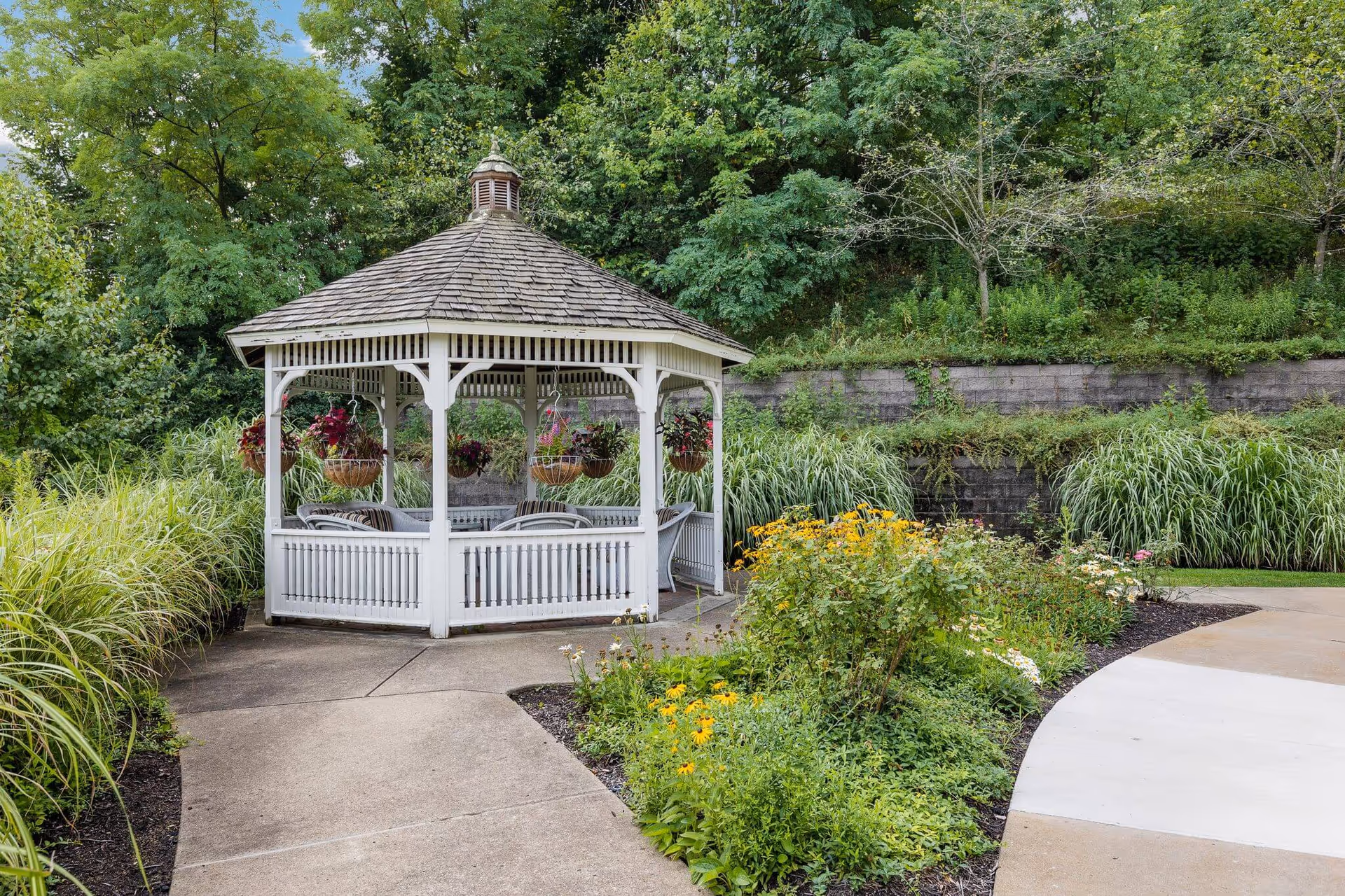 A white wooden gazebo with a shingled roof is situated in a garden area surrounded by lush greenery and flowering plants. The gazebo contains several chairs and hanging flower baskets. A concrete pathway curves around the garden leading to the gazebo.