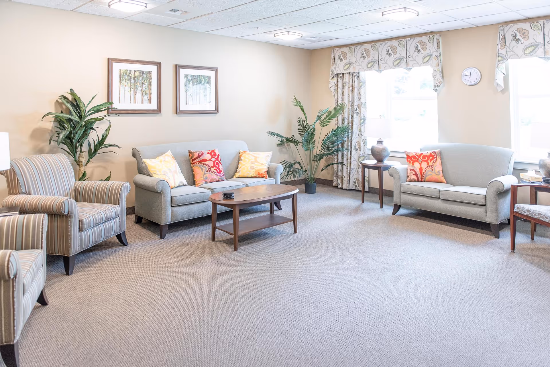 Bright communal living room with sofas, armchairs, coffee table, decorative pillows, plants, and framed art near windows.