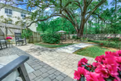 Sunny fenced courtyard with a paver patio, rocking chairs, tables with umbrellas, a large shade tree, and pink flowers in the foreground.
