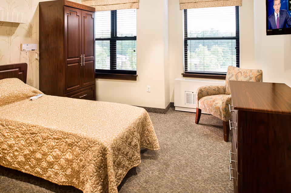 A neatly made single bed with a patterned beige bedspread in a senior living facility room. Next to the bed is a dark wooden wardrobe and a window with blinds partially open, letting in natural light. Across from the bed is a wooden dresser with a patterned armchair beside it. Another window with blinds is behind the chair, and a wall-mounted TV is visible in the upper right corner.