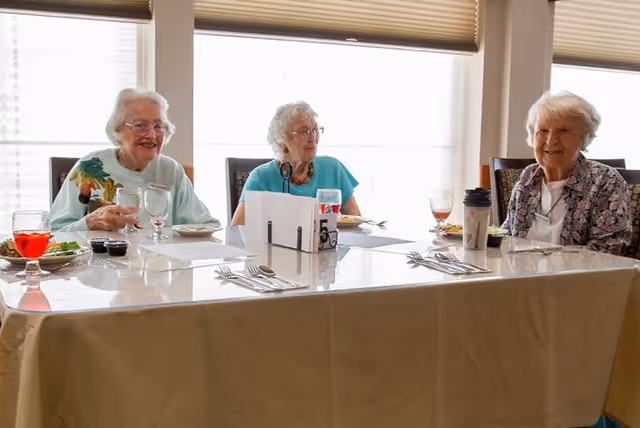 Three elderly women sitting at a dining table in a well-lit room with large windows covered by blinds. The table is set with plates, glasses, utensils, and some food and drinks. The women appear to be engaged in conversation and smiling.