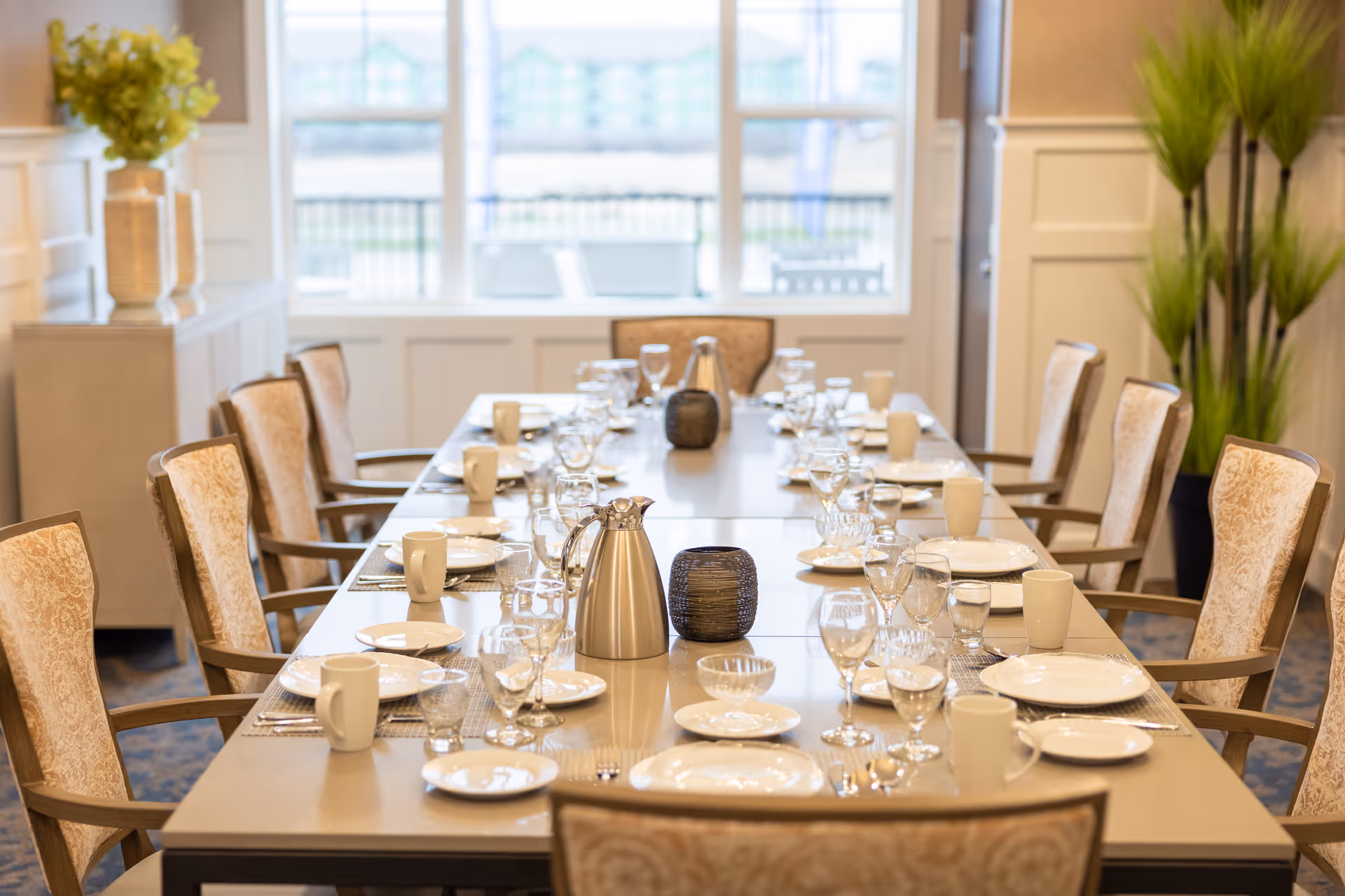 A long dining table set for a meal with plates, glasses, mugs, and silverware in a bright room with large windows and upholstered chairs. There are decorative plants and vases in the background.