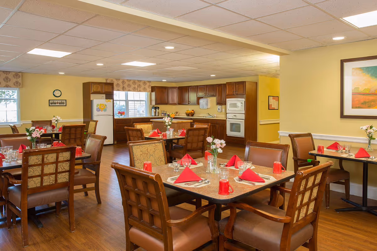 Bright dining room with multiple set tables and chairs, red napkins and flowers, and a kitchen area in the background.