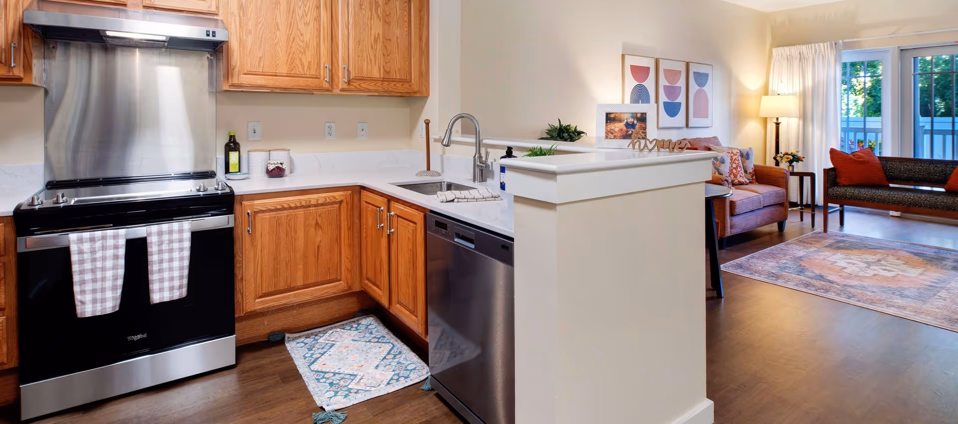 Open kitchen with stainless-steel stove and dishwasher, oak cabinets and a breakfast bar opening into a furnished living room with sofa and rug.