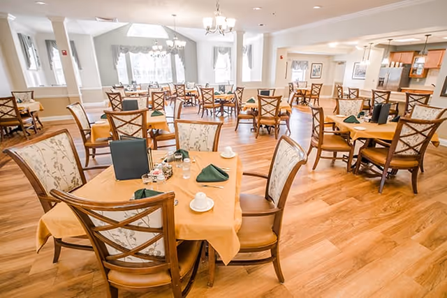 A bright and spacious dining room with multiple wooden tables and chairs arranged neatly. Each table is covered with a light orange tablecloth and set with green folded napkins, cups, and small condiment containers. Large windows allow natural light to fill the room, and chandeliers hang from the ceiling.