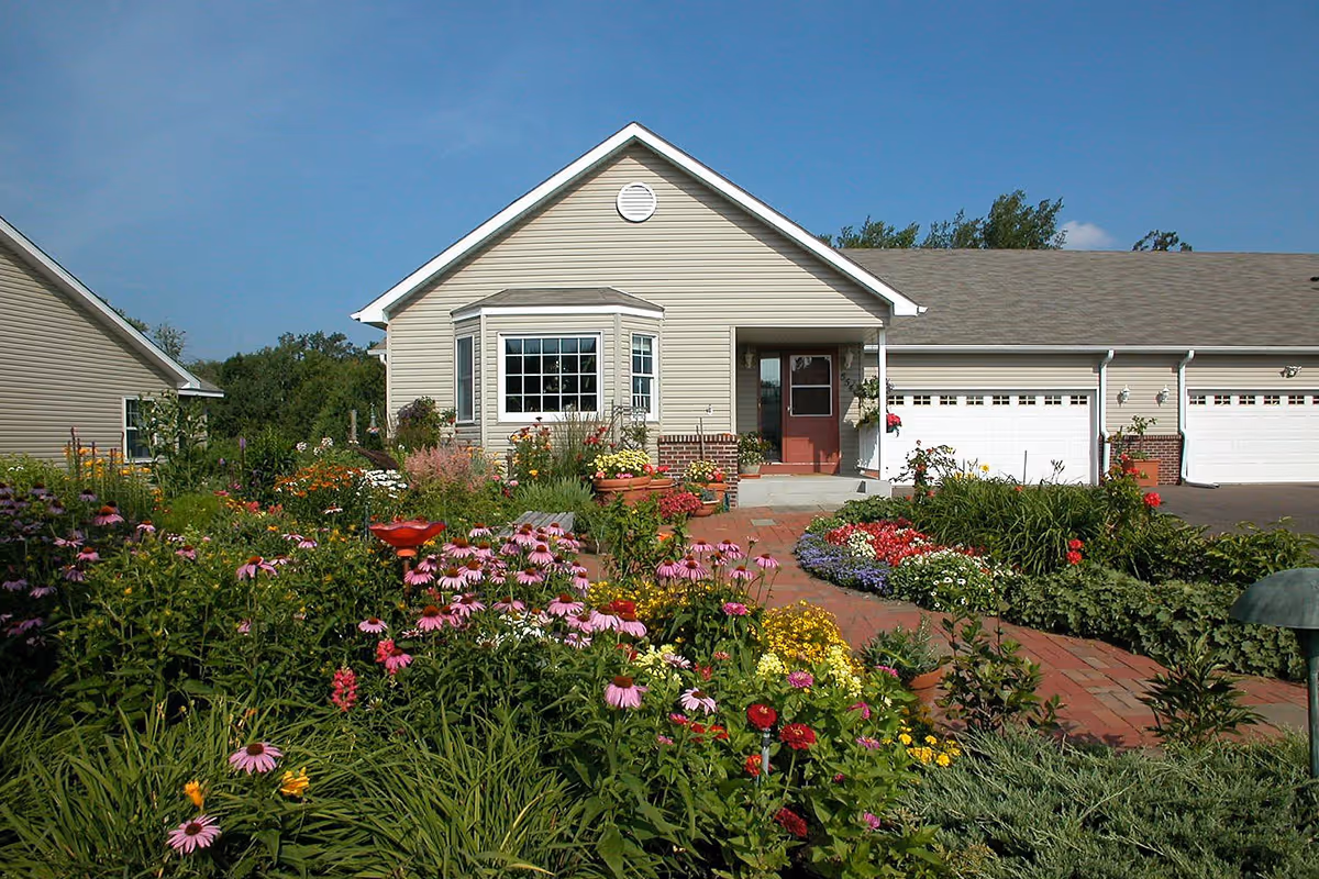 A single-story beige house with a red front door and white garage doors, surrounded by a vibrant garden filled with various colorful flowers and green plants under a clear blue sky.