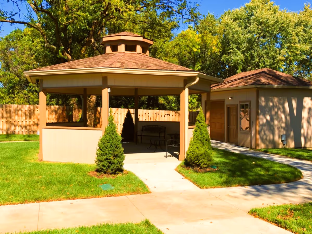 Outdoor gazebo structure with a brown shingled roof and beige walls, surrounded by green grass and small evergreen shrubs. A paved walkway leads to the gazebo, and there is a small building with doors and windows adjacent to it. Trees and a wooden fence are visible in the background under a clear blue sky.