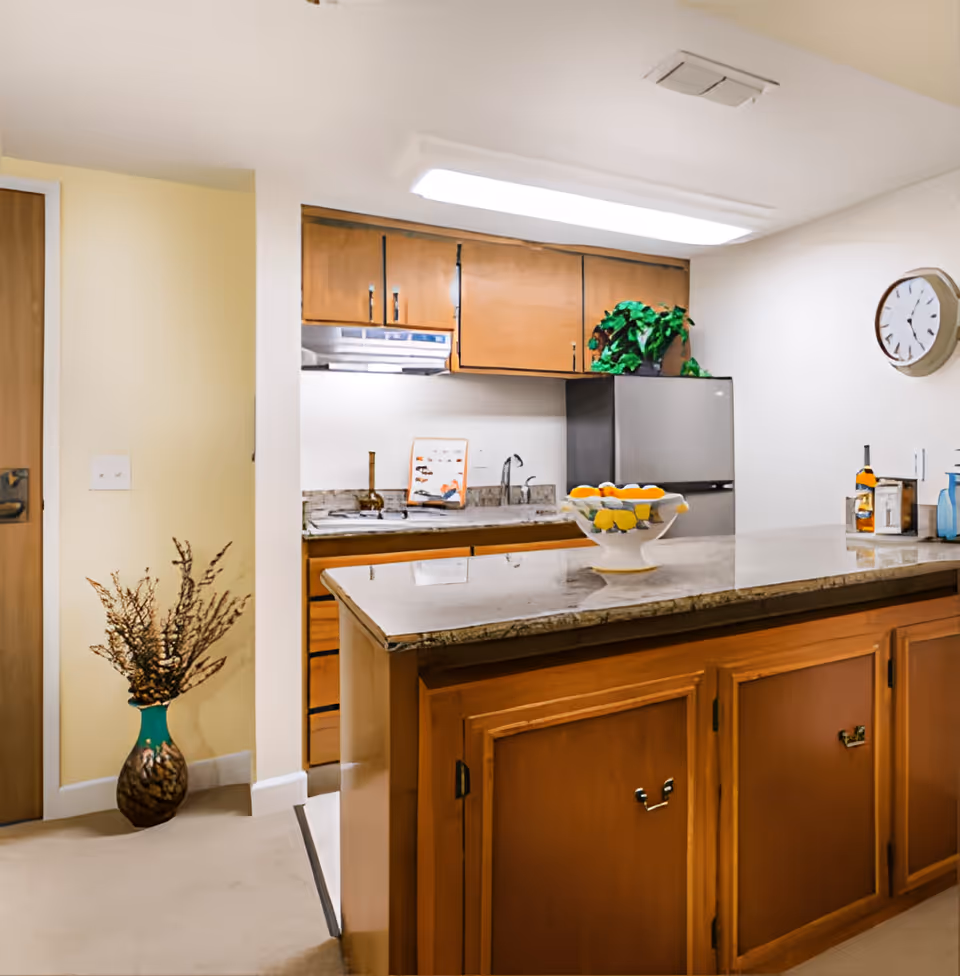 A kitchen area with wooden cabinets, a granite countertop island, a stainless steel refrigerator, a sink, and a wall clock. There is a decorative vase with dried branches on the floor and a bowl of lemons and oranges on the countertop. A potted plant is placed on top of the refrigerator.