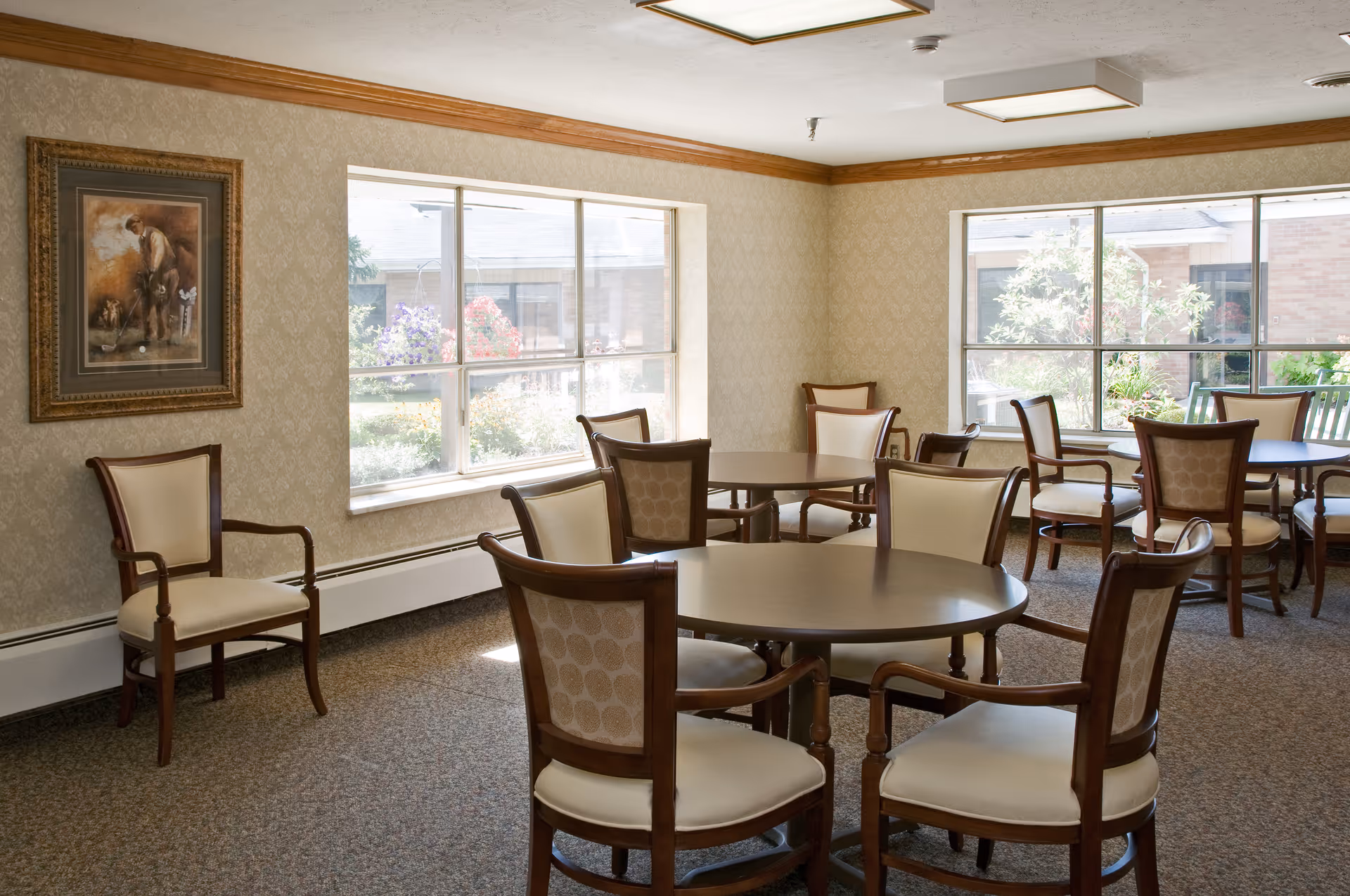 Sunlit common dining room with round tables and wooden chairs by large windows.