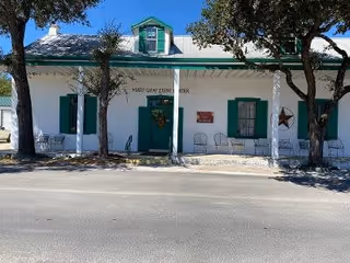 Front exterior view of a white building with green window shutters and a green door. The building has a small porch with white columns and several chairs lined up along the front. There are two trees partially shading the building and a wreath hanging on the door.