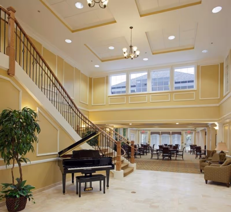 Spacious and elegant interior of a senior living facility featuring a grand piano with a bench near a staircase with wooden handrails. The room has high ceilings with recessed lighting and chandeliers, large windows letting in natural light, and a seating area with tables and chairs in the background. There is also a potted plant near the piano and comfortable armchairs to the right.