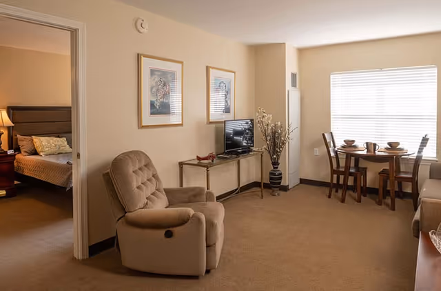 A cozy senior living apartment interior featuring a beige recliner chair, a small TV on a glass table, two framed pictures on the wall, a dining table set with four chairs and tableware near a window with blinds, and a glimpse into a bedroom with a bed and nightstand with a lamp.