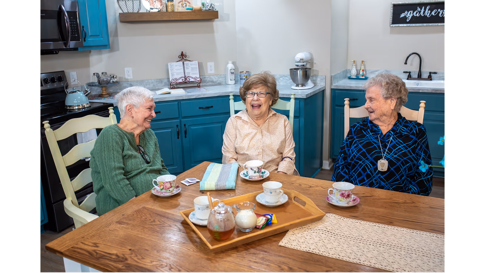 Three elderly women sitting around a wooden table in a kitchen, smiling and enjoying tea together. The kitchen has teal cabinets, a stove, a microwave, and various kitchen items on the counters.