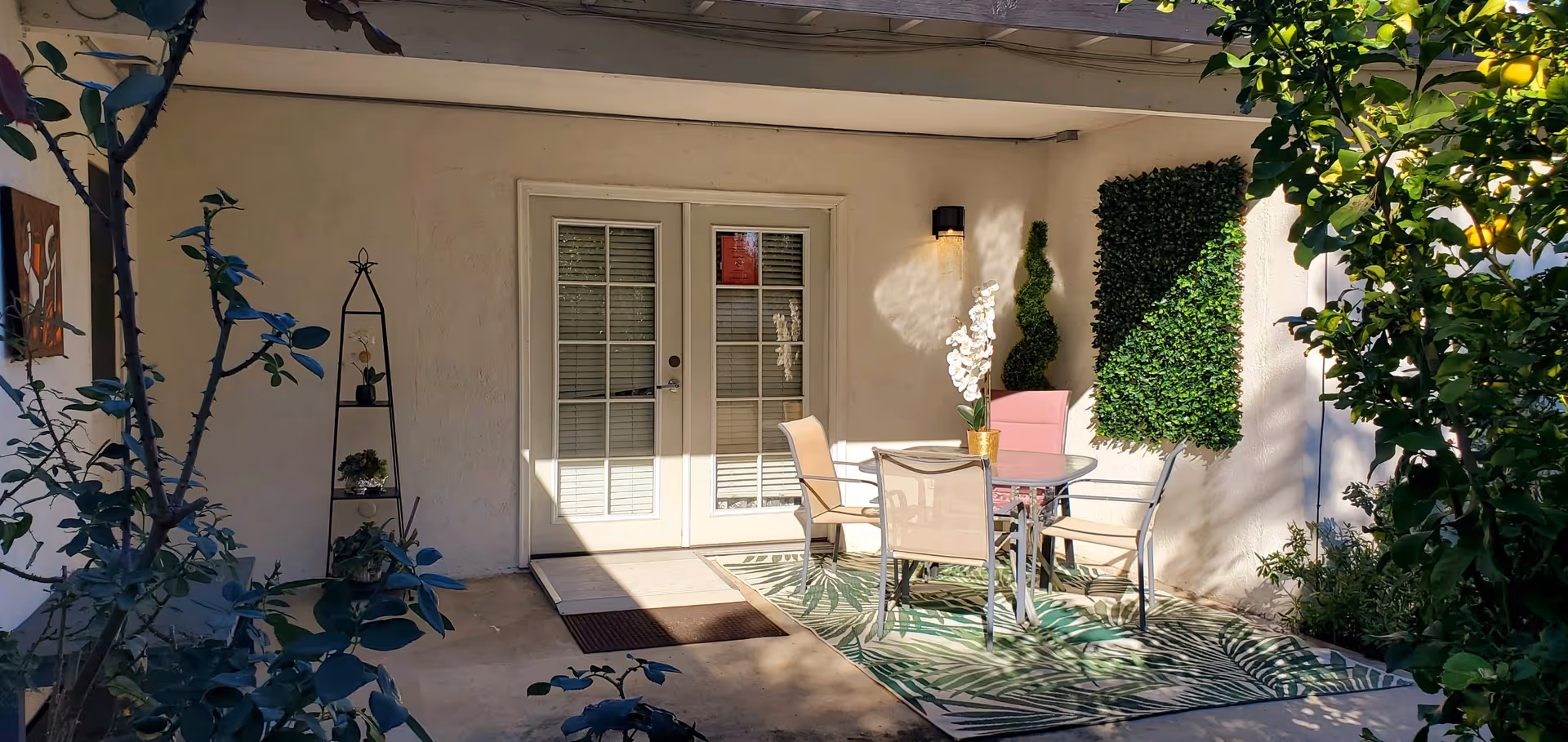 Patio area with glass doors, seating, rug, and surrounding greenery.