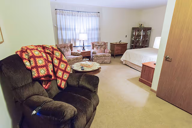 A cozy senior living room and bedroom area featuring a dark recliner with a colorful crocheted blanket draped over it, two floral upholstered armchairs with a small round table and lamp between them, a wooden dresser, a glass-front cabinet, and a bed with white bedding. The room has beige carpet and light-colored walls with a window covered by sheer curtains.