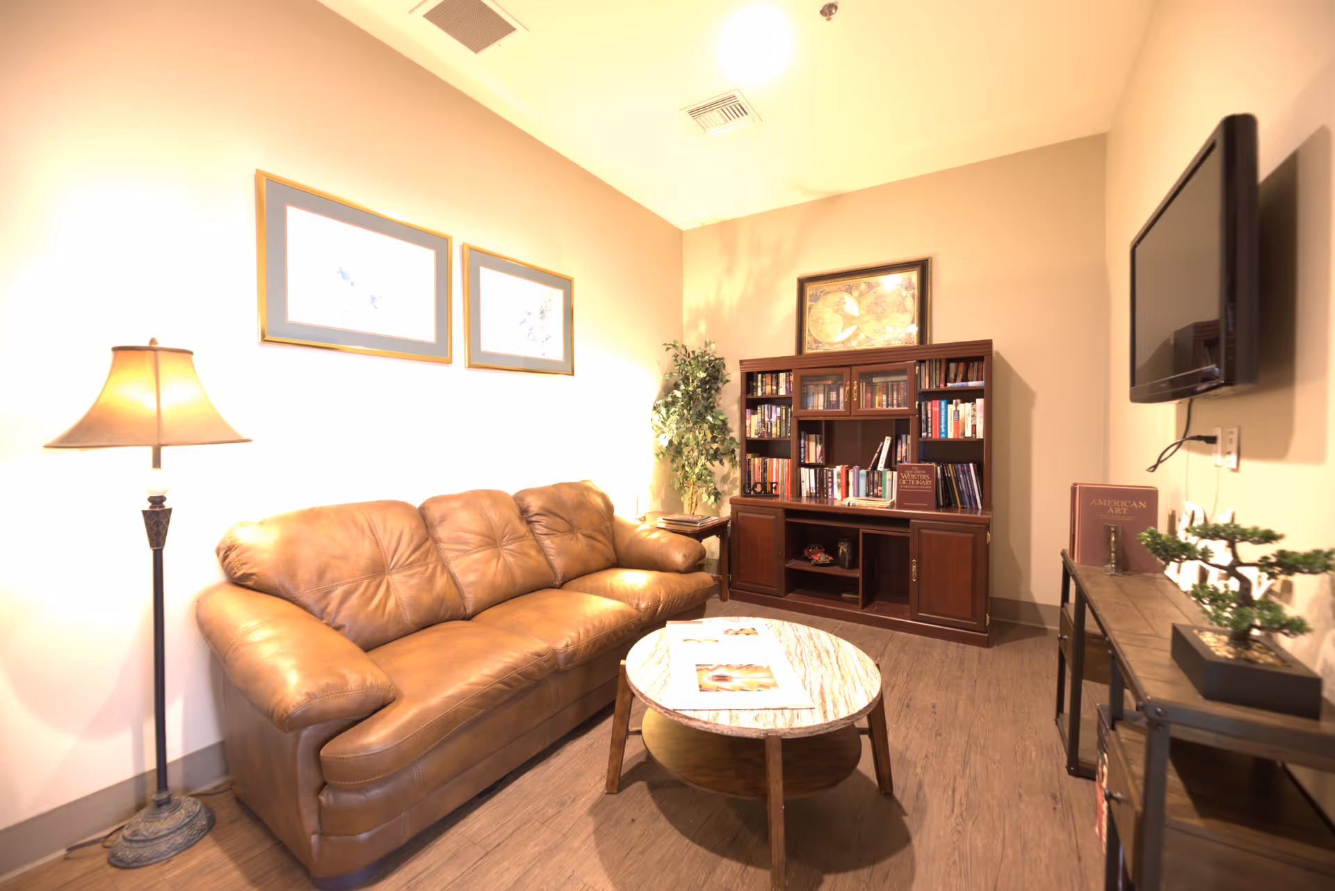 A cozy living room with a brown leather sofa, a round wooden coffee table with magazines, a tall floor lamp, a wooden bookshelf filled with books, a potted plant, a wall-mounted flat screen TV, and framed artwork on the walls.
